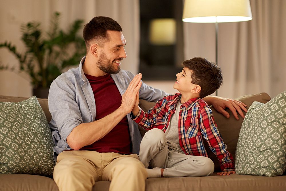 A father and his young son sitting on a sofa, mid high-five, both laughing.