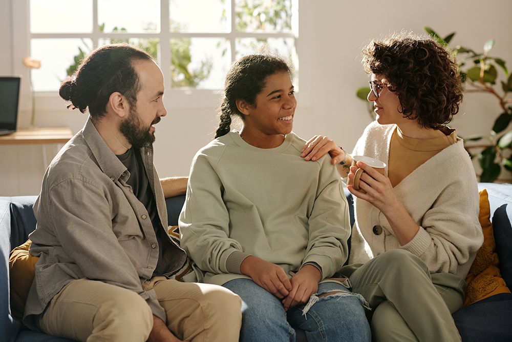 Two parents and their teenage daughter sitting close together on a sofa, talking.