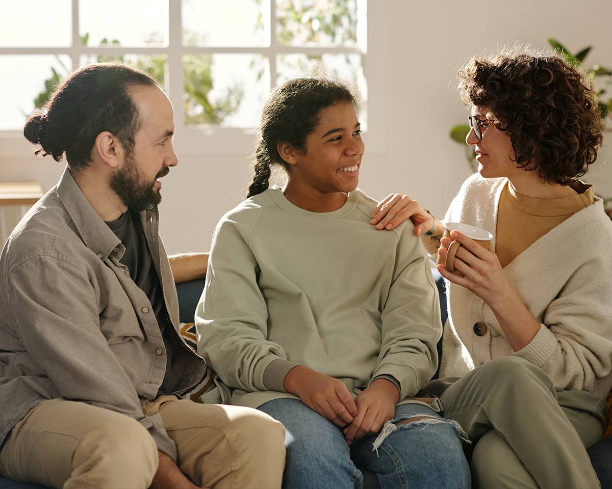 A therapist sitting with a teenager and a parent on a sofa, all three in conversation.