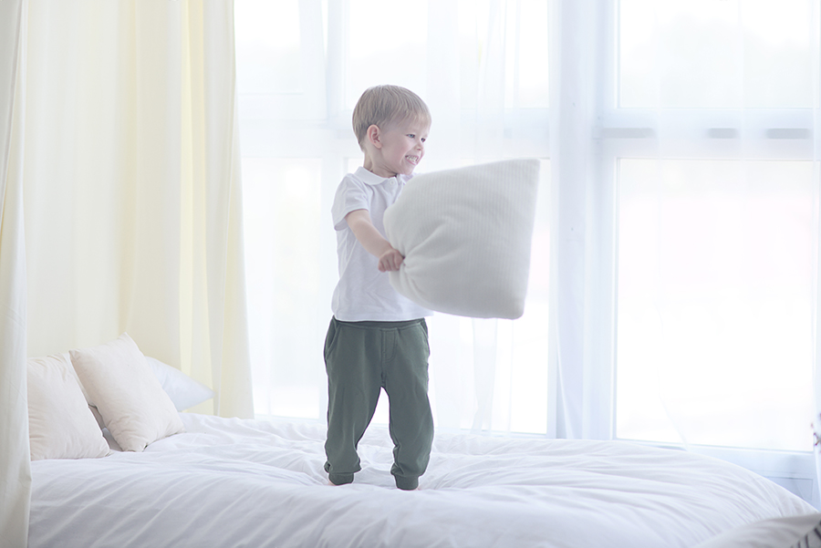 A young boy standing on a bed in a bright bedroom, swinging a pillow.