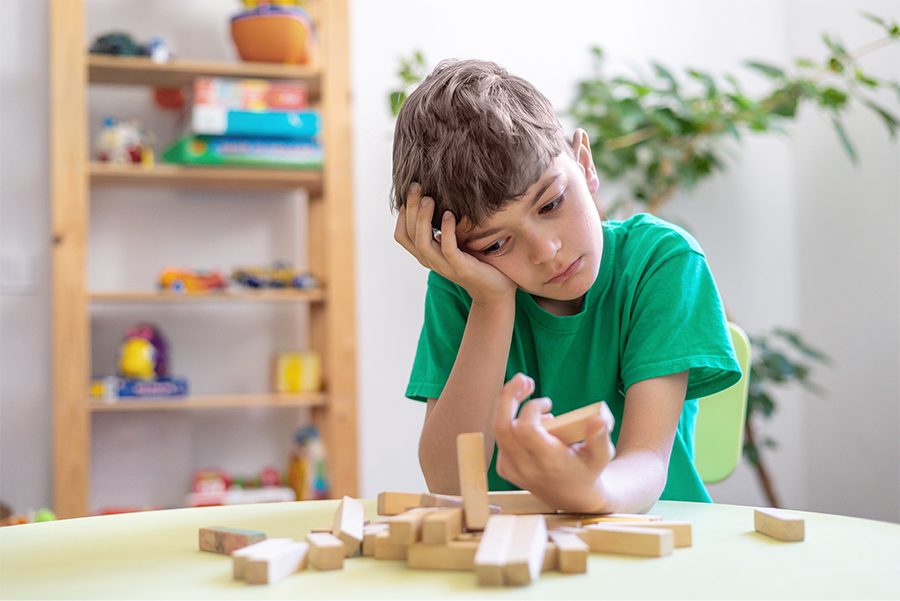 A boy resting his head on his hand at a playroom table, looking down at a scattered pile of wooden blocks.