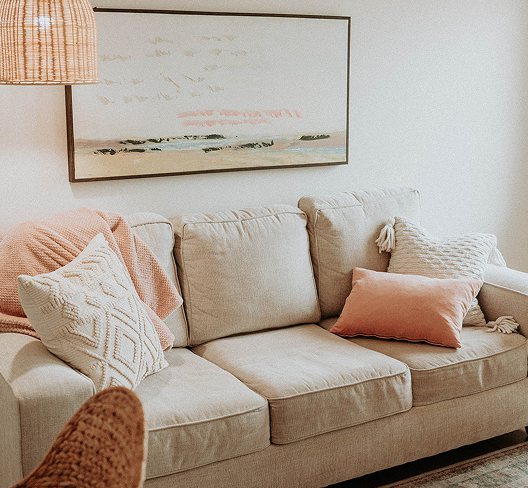 The Willow Tree waiting area: a cream sofa with coral and knitted cushions, a rattan pendant lamp, and a framed shoreline painting above.