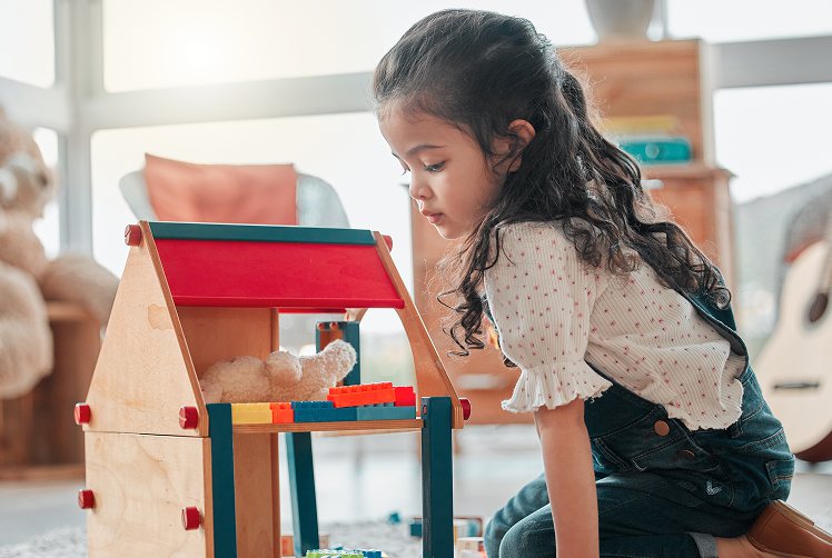A young girl kneeling on the floor beside a wooden play house, absorbed in her game.